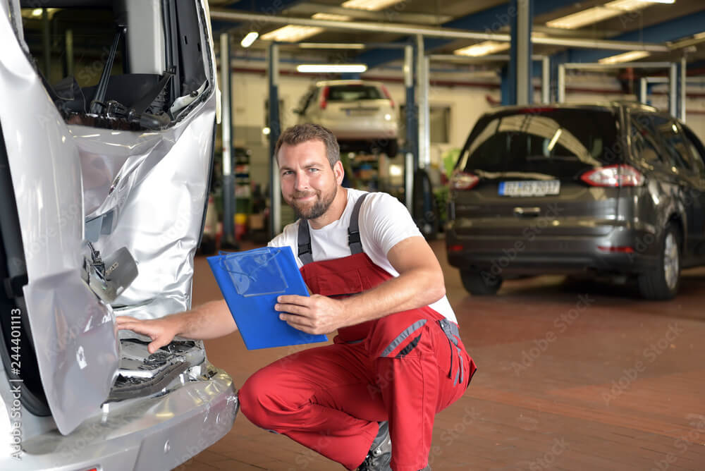 Ein Kfz-Schadengutachter in roter Arbeitskleidung prüft mit einem Klemmbrett in der Hand eine beschädigte Stoßstange in der Werkstatt