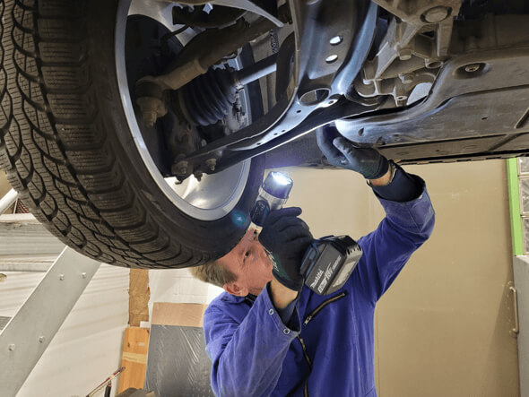 Ein Mechaniker in blauen Overalls untersucht mit einer Taschenlampe und einem Akku-Gerät die Unterseite eines Autos