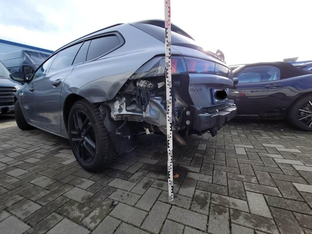 Ein graues Auto mit einem Schaden am Heck steht auf einem asphaltierten Parkplatz; neben der Beschädigung liegt ein Messstab.