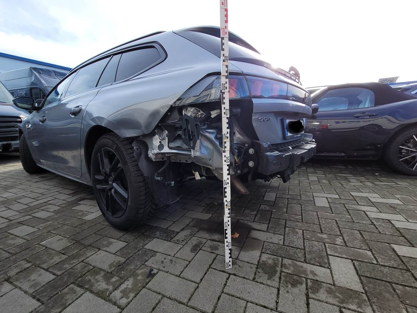 Ein graues Auto mit einem Schaden am Heck steht auf einem asphaltierten Parkplatz; neben der Beschädigung liegt ein Messstab.