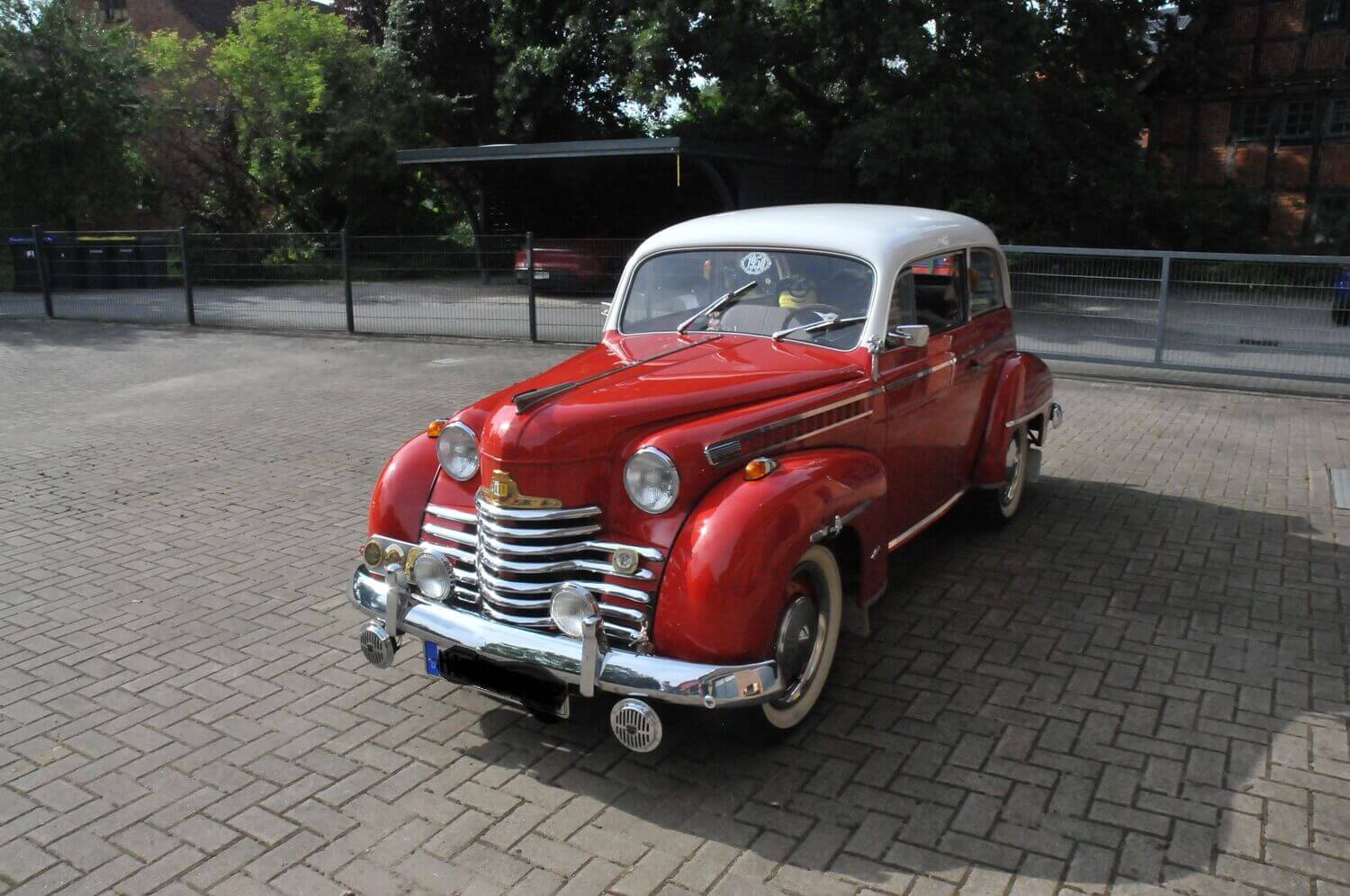 Ein glänzender rot-weißer Oldtimer steht auf einem asphaltierten Parkplatz neben einem Zaun und Bäumen