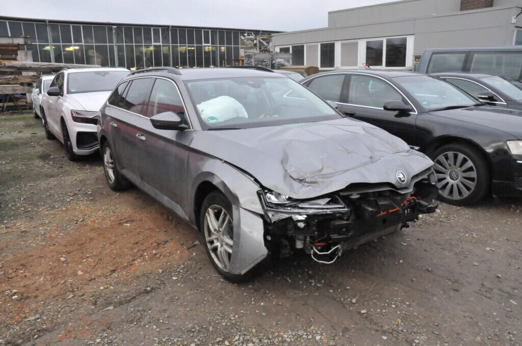Ein silbernes Auto mit beschädigter Front, fehlendem Stoßfänger und fehlenden Scheinwerfern steht auf einem Parkplatz neben anderen Fahrzeugen