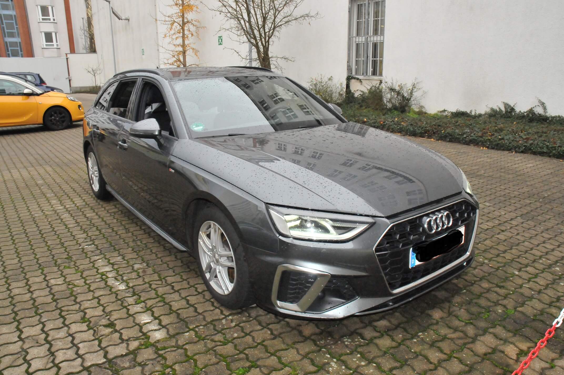 Ein grauer Audi-Kombi, der einem Kfz-Sachverständigen gehört, steht regennass auf einem gepflasterten Parkplatz neben einem weißen Gebäude