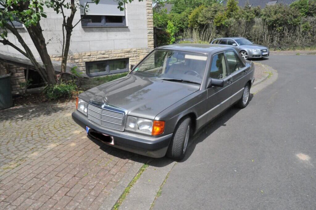 Ein grauer, klassischer Mercedes-Benz steht in einer Wohnstraße neben einem Gehweg und einem Baum