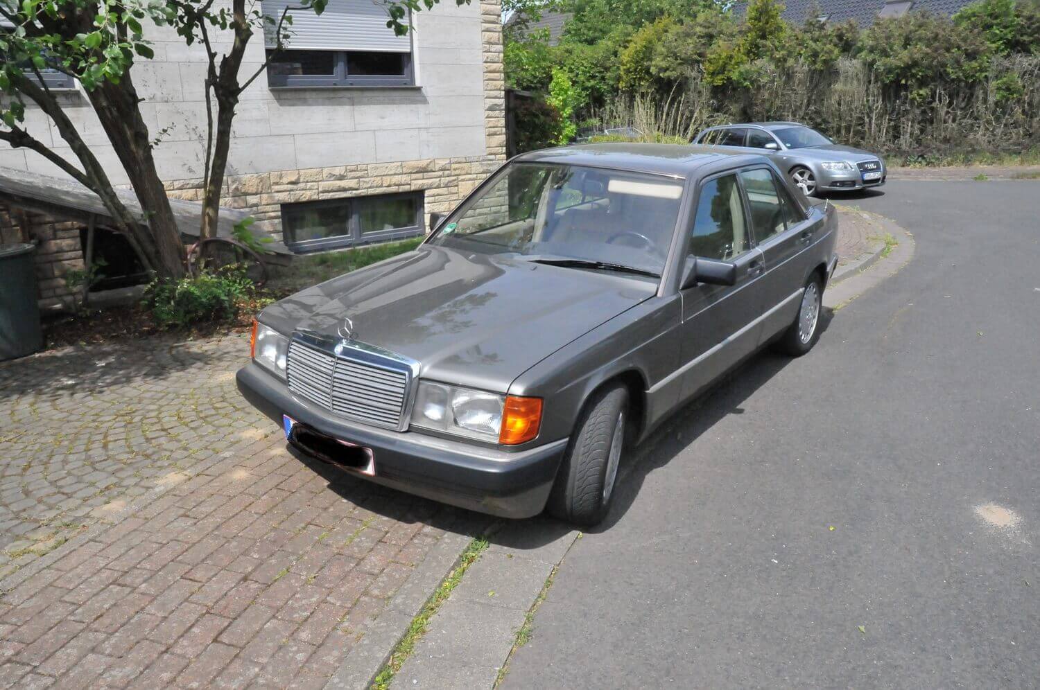 Ein grauer, klassischer Mercedes-Benz steht in einer Wohnstraße neben einem Gehweg und einem Baum