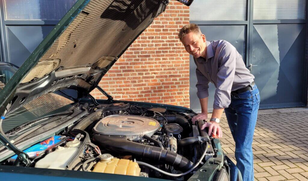 Ein Mann in blauen Jeans und einem grauen T-Shirt lächelt, während er draußen vor einer Backsteinmauer neben einem Auto mit offenem Motor steht