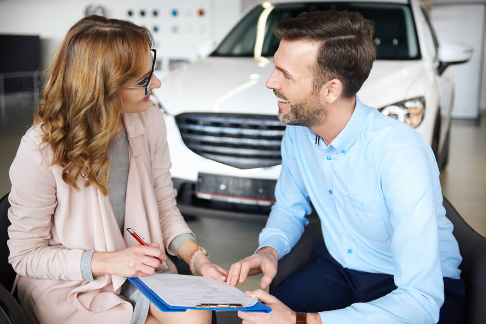 Eine Frau und ein Mann besprechen Unterlagen zum Marktwert vor einem weißen Auto im Autohaus